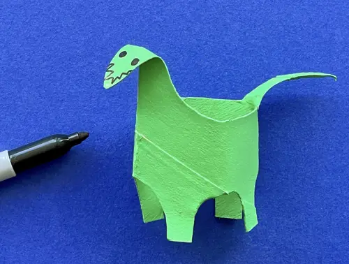 Een groene dino van wc-rol waarop oogjes en tanden zijn getekend met de zwarte viltstift die ernaast ligt.
