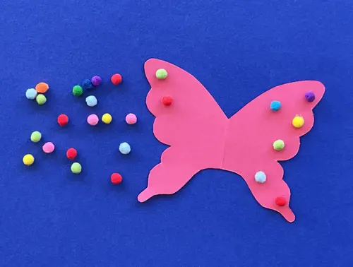 Roze kartonnen vlinder waarop kleine gekleurde bolletjes zijn geplakt; er liggen meer van de bolletjes naast.