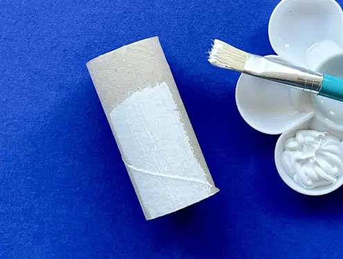 Half geverfde kartonnen wc-rolkokertje op een blauwe achtergrond naast een penseel en witte verf.