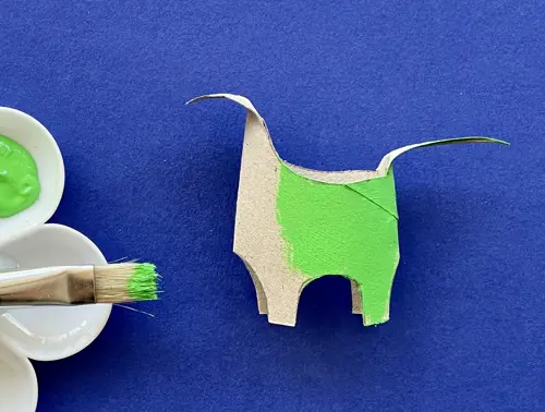 Een deels groen geverfde dino van wc-rol met een kunststof palet met groene verf en een penseel ernaast.